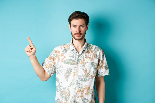 脸度假时留着胡子的帅哥的肖像 穿着夏装 用手指着商标 站在蓝色的背景上男性胡子帅哥