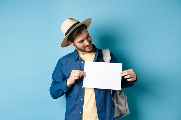 男快乐的游客戴着草帽微笑着看着一张纸为你的标志 站在蓝色的背景上酷男成人