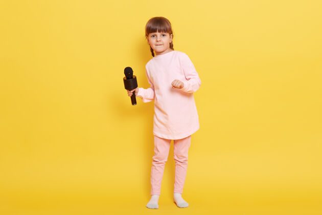 麦克风迷人的小女孩 带着麦克风 穿着粉色浅色的衣服 孤立地站在黄色背景下 迷人的歌手在卡拉OK里唱歌 孩子们在安排音乐会声音人人