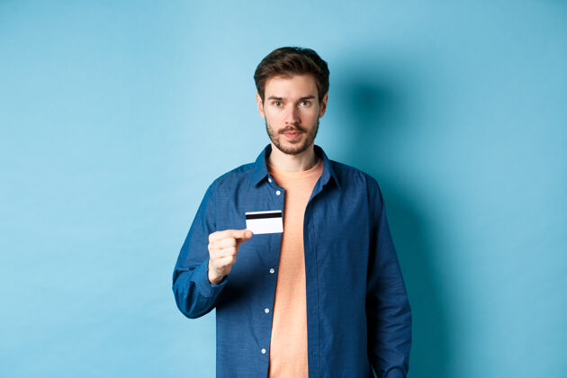 购物者帅哥留着胡子 穿着休闲服 拿着塑料信用卡 看着相机 蓝色背景时尚情感购物