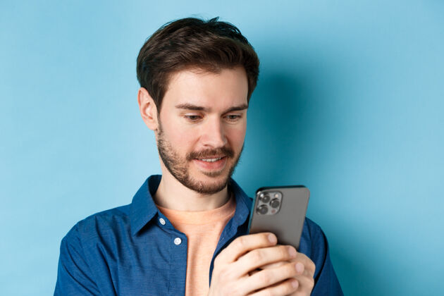 帅气一个快乐的年轻人的特写照片 他正在读手机 在智能手机里联网 站在蓝色的背景上休闲年轻时尚