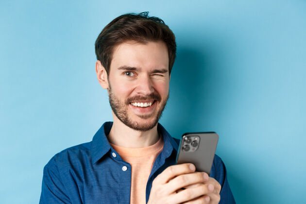 聊天开朗的男人眨眼微笑的特写镜头 在蓝色背景下使用手机年轻帅气手机