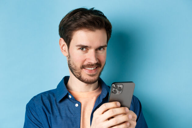 成人特写镜头中 一个迷人的白人男子手持手机 对着镜头微笑 穿着休闲装站在蓝色背景下社交网络酷