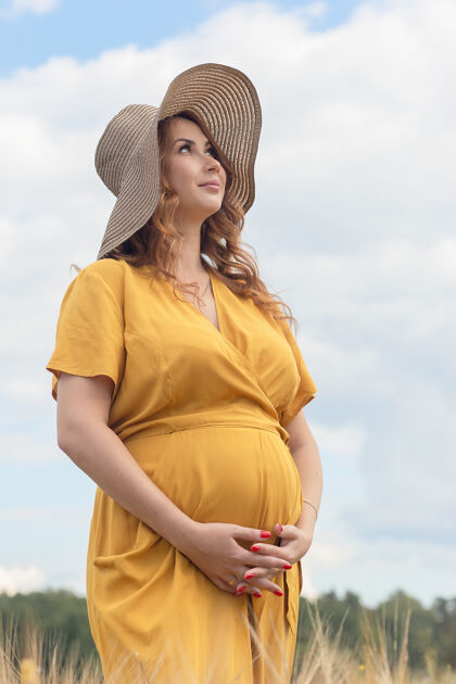 太阳在一个阳光明媚的夏日 一位年轻漂亮的孕妇穿着黄色的裙子 戴着黄色的帽子 走过一片麦田收获农村帽子