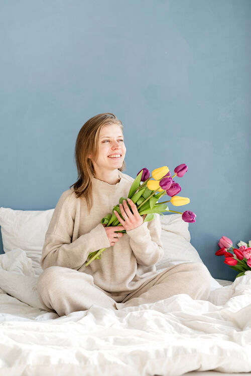 快乐快乐的女人穿着睡衣坐在床上捧着郁金香花束 背景是蓝色的墙壁郁金香女性卧室