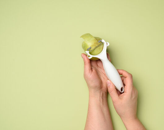 餐具女性的手拿着一把清洗蔬菜水果的塑料刀和一个绿色背景上的绿色苹果 特写厨具锋利切割
