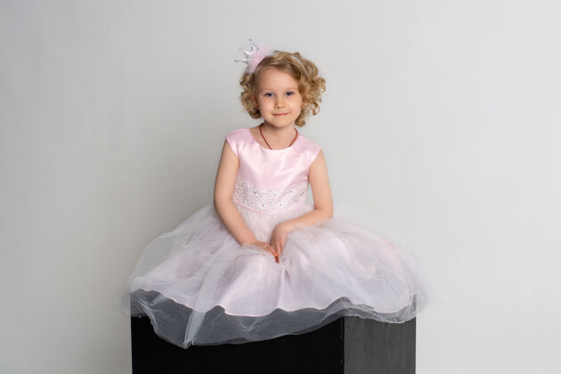 有趣一个小女孩穿着粉红色的裙子 头戴皇冠 坐在白色背景的黑色立方体上可爱女性欢呼