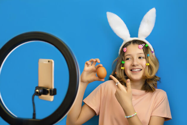 兔子开朗的少女博客作者头上戴着兔子耳朵 手里拿着一个鸡蛋 用智能手机进行在线广播手机女孩人
