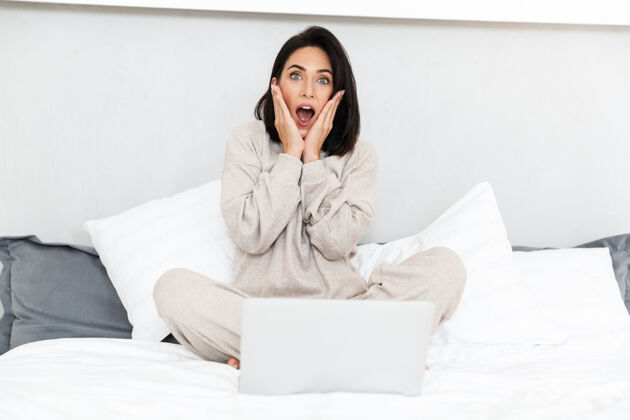 满足一张30多岁的震惊女人在舒适的公寓里用笔记本电脑 穿着白色亚麻布坐在床上的照片舒适肖像室内