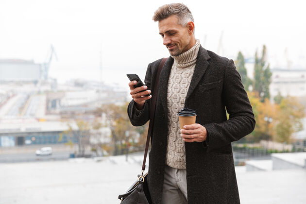 手持一张30多岁的留胡子男人的照片 他穿着夹克 拿着外卖咖啡 一边用手机 一边走在城市街道上成人肖像站立