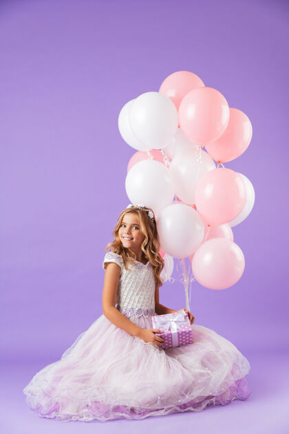 欢乐穿着公主裙的漂亮小女孩孤零零地坐在紫罗兰色的墙上 手里拿着一堆气球和一个礼盒微笑白种人气球