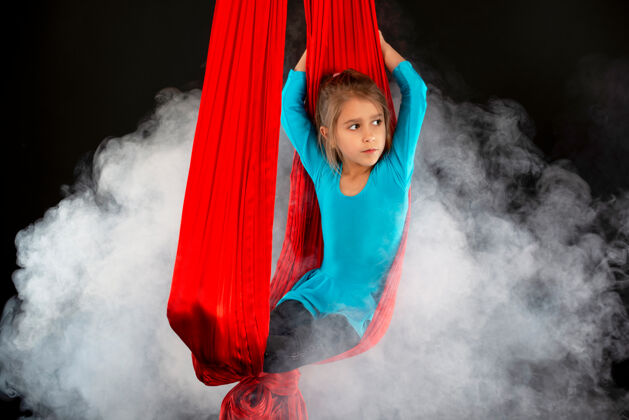 健身房身穿蓝色体操服的无畏美丽小女孩在黑色的舞台上表演特技空中红丝带 周围烟雾缭绕美容戒指私人教练