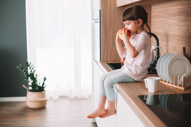 生活方式坐在厨房台面上吃红苹果的可爱小女孩健康饮食理念健康水果独自