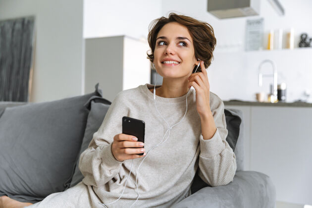 耳机快乐的年轻女子坐在家里的沙发上 用手机 用耳机听音乐音频声音女士