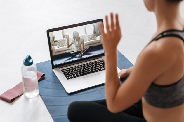 电脑穿着运动服的年轻健身女教练用笔记本电脑在视频会议上进行在线健身训练或虚拟瑜伽课的特写镜头热身平衡地板