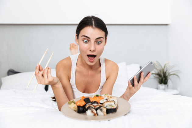 吸引美丽的年轻女子在家里的床上放松 拿着手机 吃着盘子里的寿司床室内青年