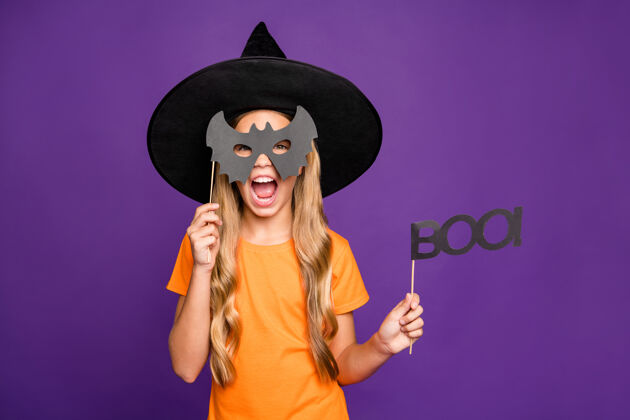 万圣节小巫婆扮演超自然角色的照片万圣节主题派对手持蝙蝠纸棒恐怖造型穿橙色t恤巫师帽孤立紫色背景邪恶年轻未成年