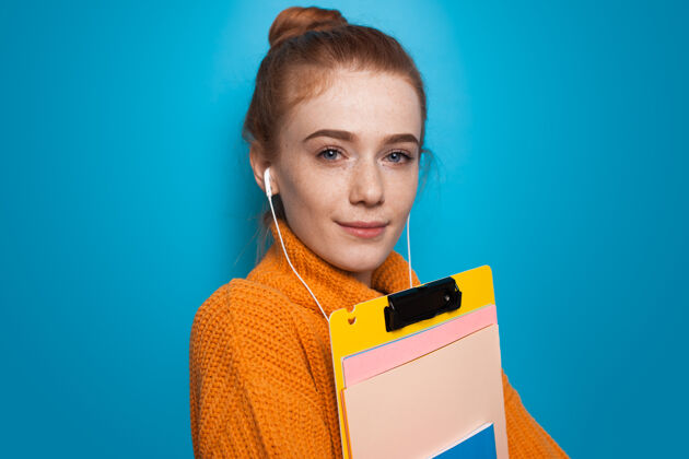 文件夹一个有雀斑的姜学生穿着黄色毛衣 拿着文件夹和耳机在蓝色工作室墙上摆姿势的特写照片微笑欧洲水平