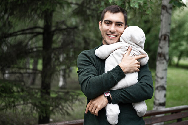 父亲节带着微笑的棕发父亲 穿着绿色夹克 怀里抱着孩子 在公园里散步 还有复印空间关系新生儿快乐