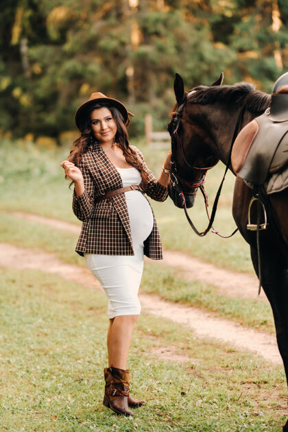 女孩一个大肚子的怀孕女孩戴着帽子在森林里的马旁边自然时尚一个穿着棕色裙子的孕妇和马在一起户外时尚女人
