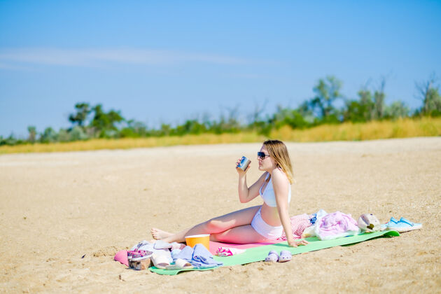 魅力阳光明媚的沙滩上 一个穿着白色泳衣的小女孩坐在垫子上 手里拿着一个杯子 喝着饮料女人马克杯年轻