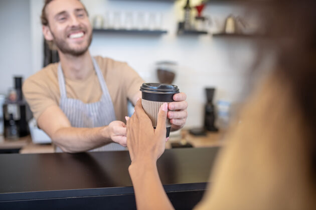 千禧一代准备就绪 咖啡微笑围裙里的年轻人把一杯咖啡递给女人的手 积极的一刻订单腰向上转移