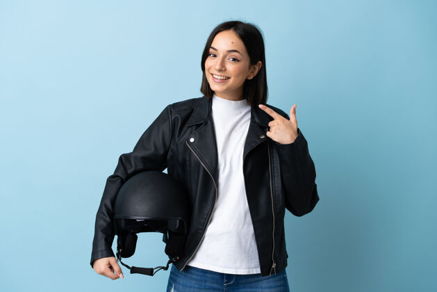 成人一个女人拿着摩托车头盔站在蓝色的墙上竖起大拇指年轻女性支架