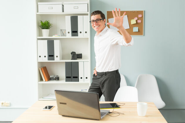 手戴眼镜的帅哥站在办公室的电脑和书架旁边专业优雅金融