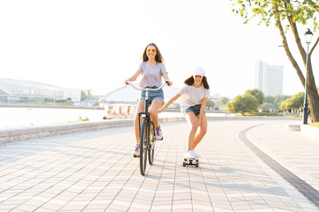 拉两个年轻的朋友在一个炎热的夏日放松 一个年轻的女人骑着自行车拖着她的朋友在一条城市街道上滑滑板友谊街道朋友
