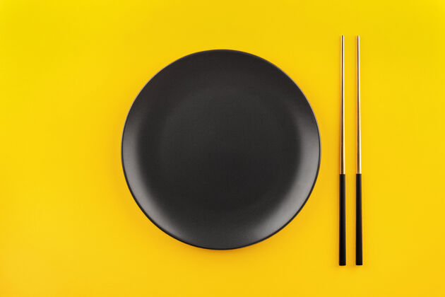 餐具用镀金筷子制作的黑色圆形空盘子平放在黄色背景上 留有复印空间圆圈盘子黄色背景