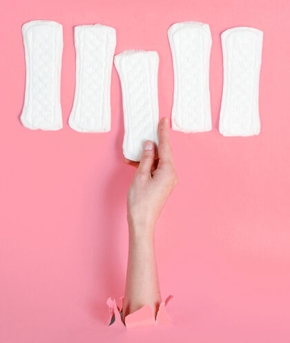 棉女性手拿卫生巾穿过撕破的粉色纸背景：极简主义时尚理念周期手臂月经