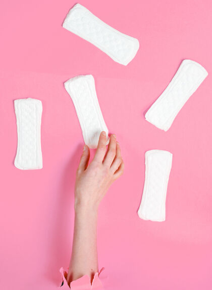 女性女性手拿卫生巾穿过撕破的粉色纸背景：极简主义时尚理念月撕裂产品