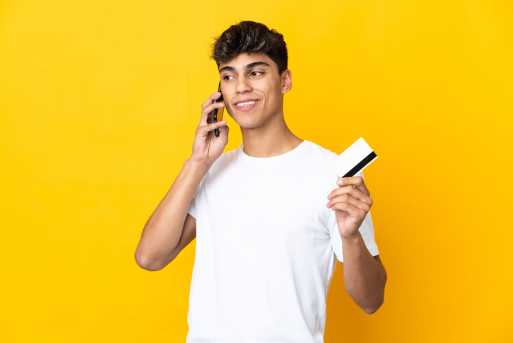 电话一个年轻人在孤立的黄色背景下拿着一张信用卡和手机交谈电话支付技术
