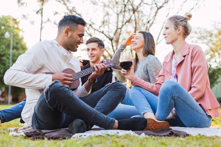 休闲一群朋友坐在草地上享受户外的乐趣——一个年轻人在演奏四弦琴 而他的朋友们则在欣赏他们的音乐和美酒假日肖像团体