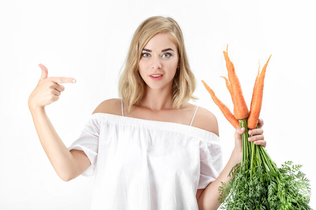 女人一位穿着白色上衣的金发美女在一张白纸上展示了一个绿叶的新鲜胡萝卜背景.健康还有节食女性成人素食