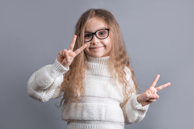 手势一个年轻迷人的金发小女孩的肖像 穿着毛衣 在灰色的工作室背景上 双手都有v字标志 和平标志 胜利手势手指头发金发