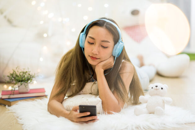 女士漂亮迷人的亚洲年轻女子在客厅用智能手机和耳机播放社交媒体和在线流媒体音乐放轻松在周末快乐年轻乐趣
