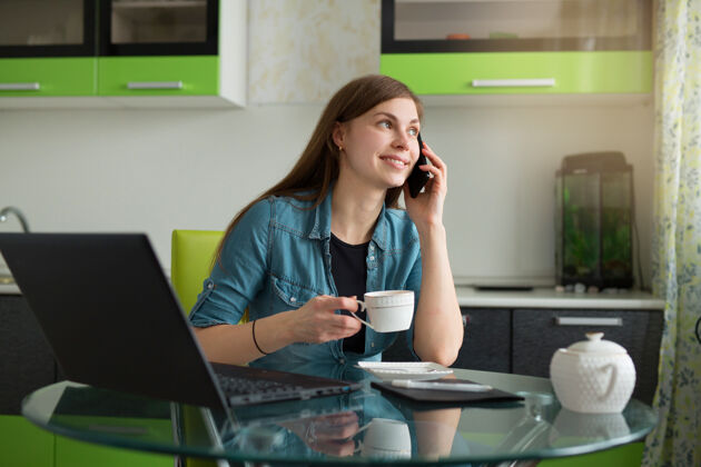 工人漂亮的年轻女子坐在厨房的桌子旁 拿着笔记本电脑喝茶 讲着电话成功连接爱好