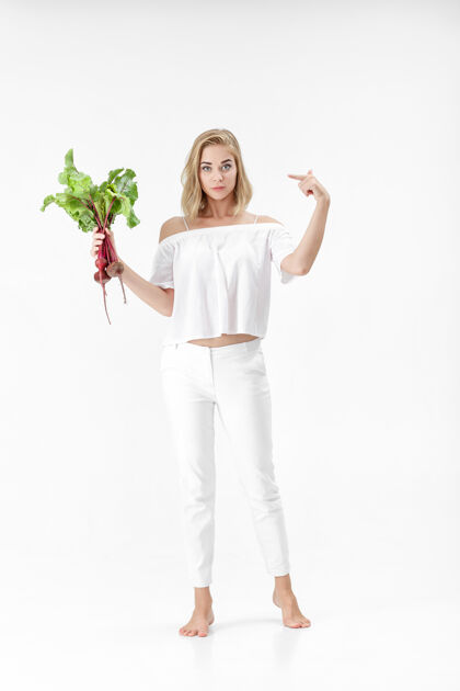 成人一位穿着白色上衣的金发美女指着一棵长着绿叶的甜菜根背景.健康还有维生素营养欢呼红色