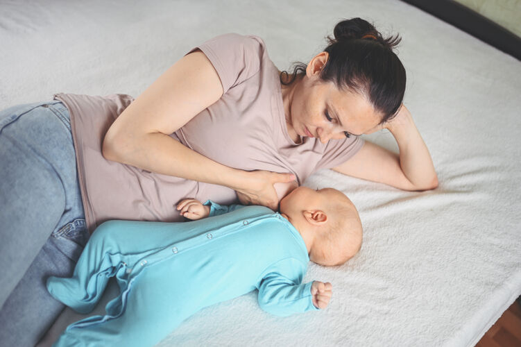 健康年轻的母亲躺在床上 抱着一个穿着蓝色连体衣的可爱的新生婴儿 用母乳给他喂奶可爱食物男孩
