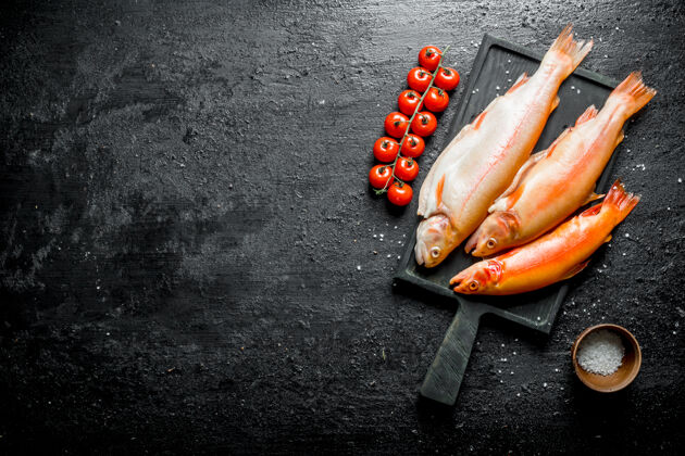自然切菜板上的生鳟鱼 树枝上放着西红柿 盘子里放着盐碗开黑色乡村背景晚餐烹饪地中海