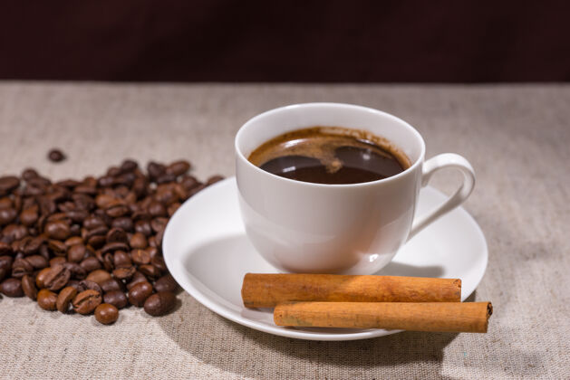 干一整杯黑咖啡 放在白色茶杯和茶碟里 背景是肉桂棒和豆子 放在桌布上分散过滤器刺激
