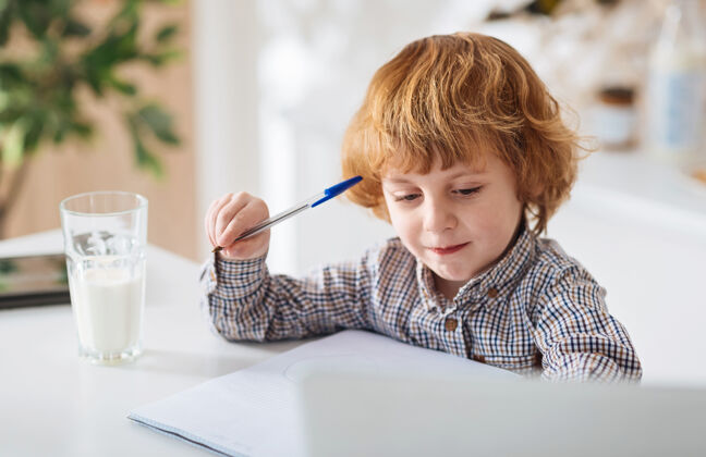 阳光好奇心是关键钥匙可爱可爱的聪明孩子坐在厨房的桌子旁 一边喝着一杯牛奶一边写着笔记学习写作勤奋