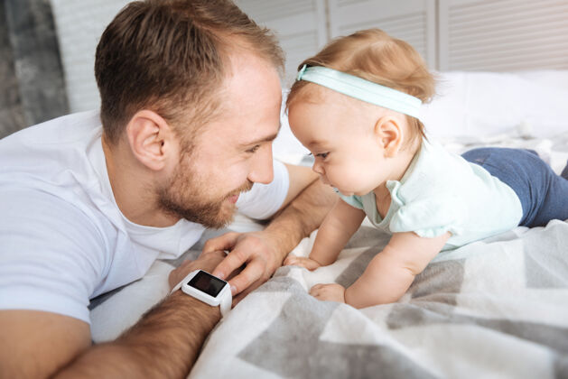 可爱享受免费时间活泼可爱的小女婴躺在床上与她的父亲 看着智能手表 同时表示兴趣和喜悦新生儿孩子互动