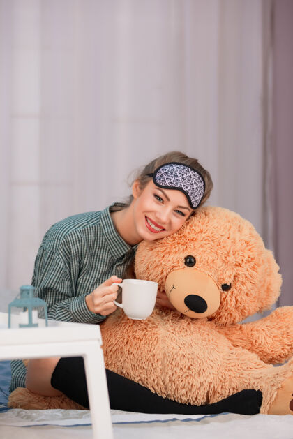 白杯子早餐床年轻女人微笑 喝咖啡茶 拥抱大玩具熊蓝眼睛放松玩具熊