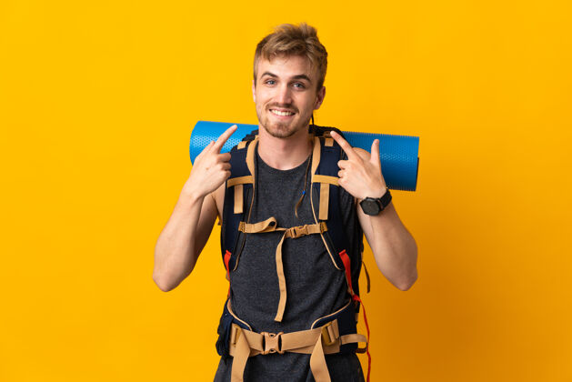 旅游者年轻的登山男子背着一个大背包 孤立地站在黄色的地面上竖起大拇指人男性旅游