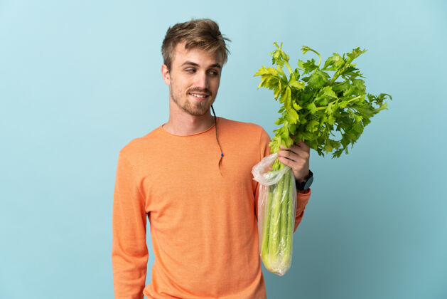 青年年轻的金发男子拿着芹菜孤立在蓝色与快乐的表情农业帅哥生的
