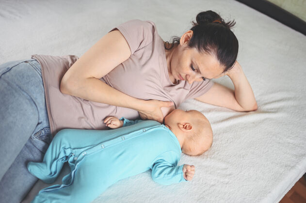 健康年轻的母亲躺在床上 抱着一个穿着蓝色连体衣的可爱的新生婴儿 用母乳给他喂奶可爱食物男孩