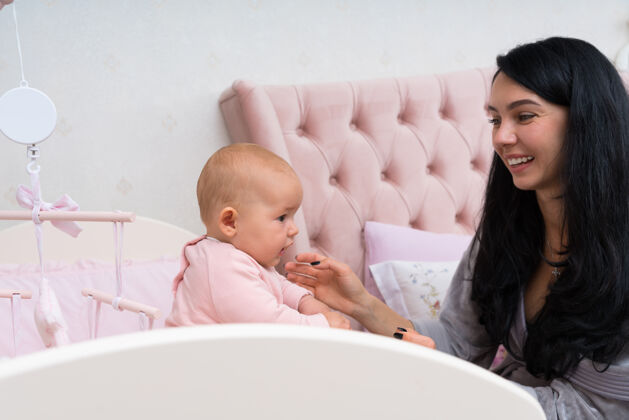 女儿一个快乐的 充满爱心的母亲和她的婴儿在粉红色的婴儿床上玩 上面挂着手机发展婴儿封闭