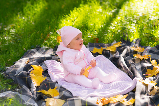 玩耍在阳光明媚的秋天公园里 小女婴坐在草地上的毯子上 草地上散落着黄色的叶子枫树分散生活方式
